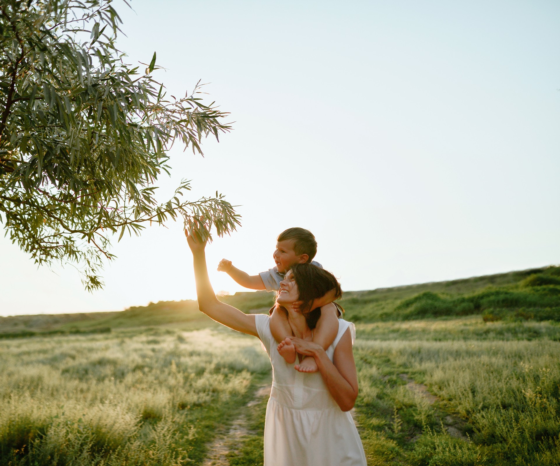 Woman carrying a toddler on her shoulders reaches up for the branch of a tree    	