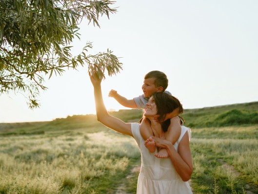 Woman carrying a toddler on her shoulders reaches up for the branch of a tree    	