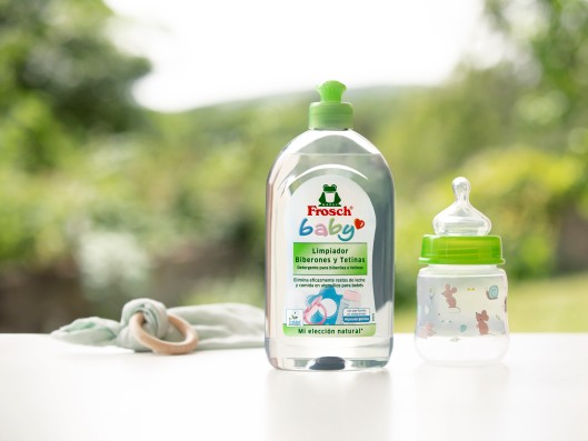 A Frosch baby dishwashing liquid bottle stands next to a baby bottle in front of a window	
